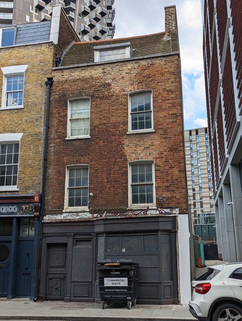 A small eighteenth century red brick house with a grey painted windows. A very tall apartment block can be seen in the background.