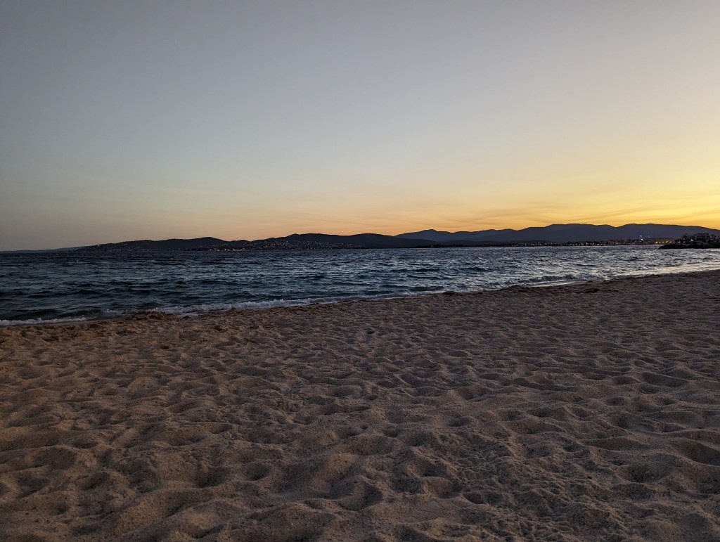 Sunset on the beach. Sand in the foreground and the grey sea behind. The sky has shades of yellow and in the distance the silhouette of Le Massif des Maures stands black against the sky.