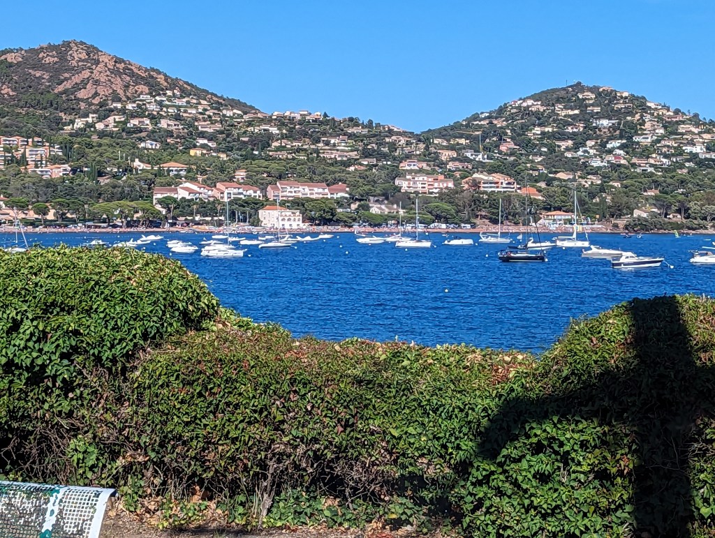 The bay of Agay with azure sea, tree lined slopes surrounding the bay, the red-brown Esterel rising above. A few small white dinghies on the water. The sky is bright blue above.