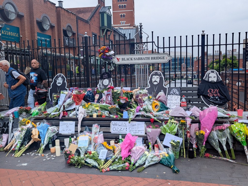 A bridge and bench in front with four images of the members of the band Black Sabbath. There are flowers covering the bench and floor in front