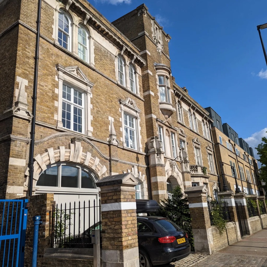 The Bethnal Green Infirmary building. A brick building with railings and pillars around it, now used as housing. The sun is shining and the sky is blue.