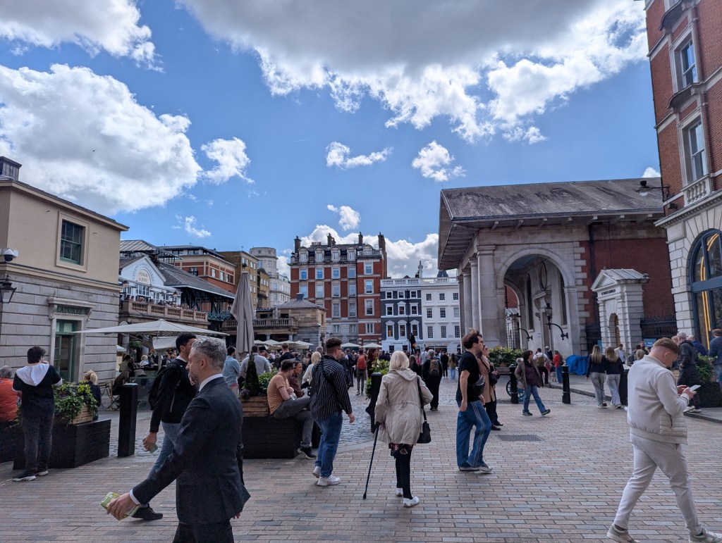 Covent Garden Piazza. The sun is shining, with a blue sky and a few clouds. People are walking about, enjoying shopping in the sunny weather.