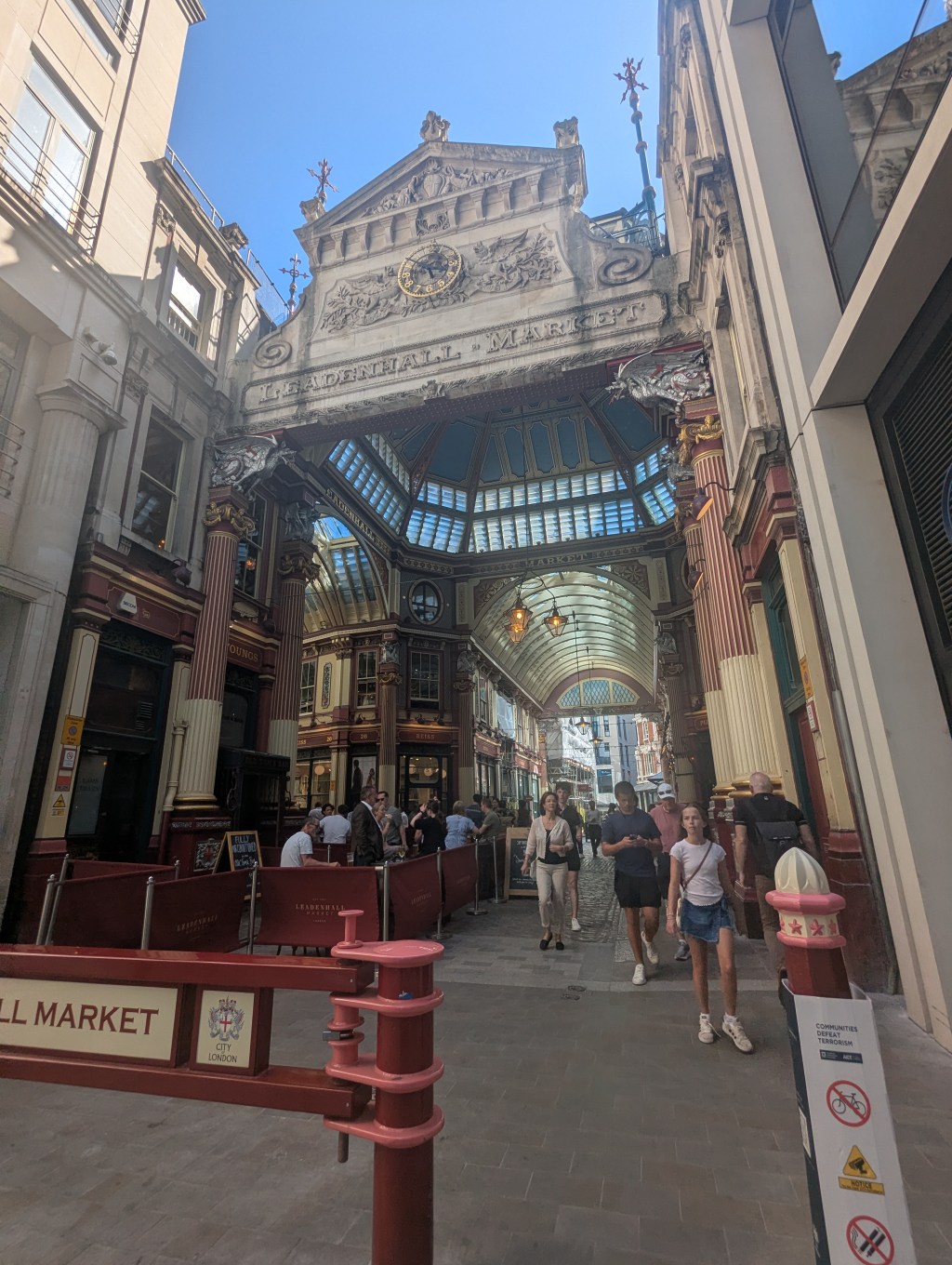 The entrance to Leadenhall Market which is a grand building with an iron and glass roof, ornate iron supports and individual shops including bars, cafes, and retail shops. It is a sunny day and the sky is blue.