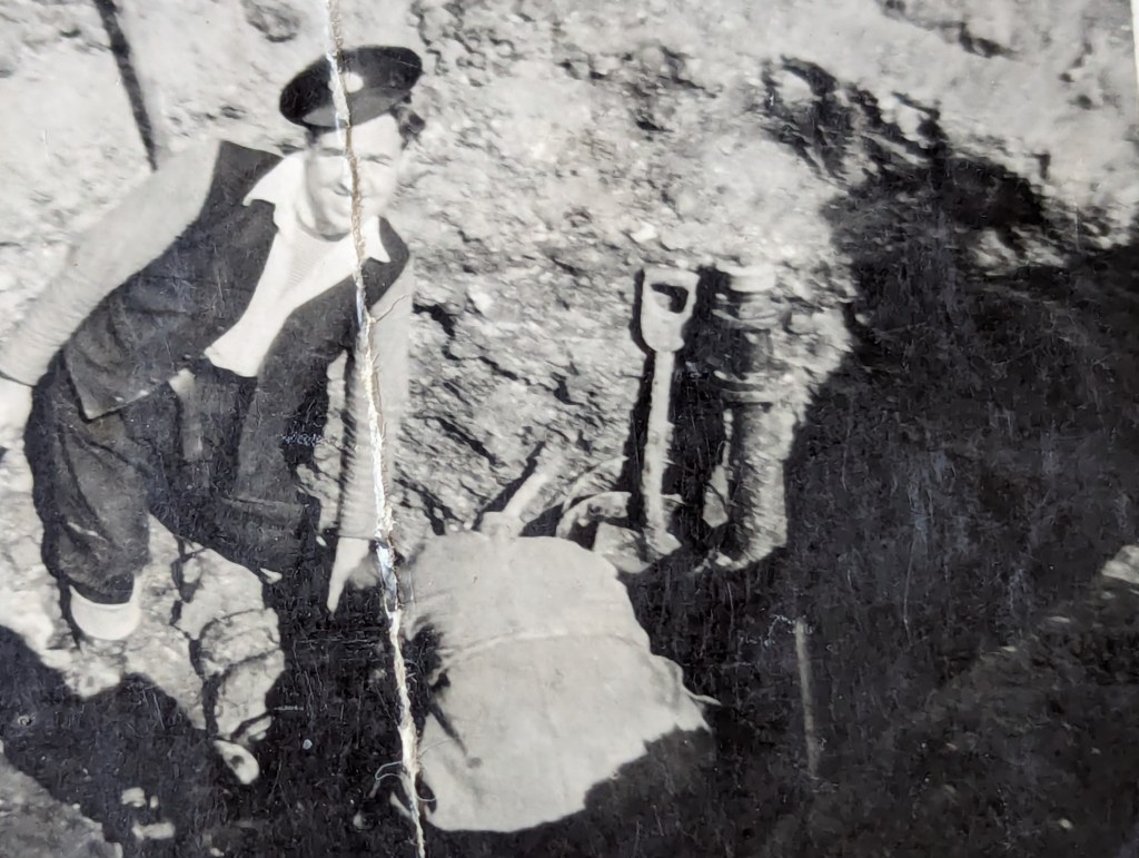 A black and white photograph showing a man working in a trench in a road repairing a water main.