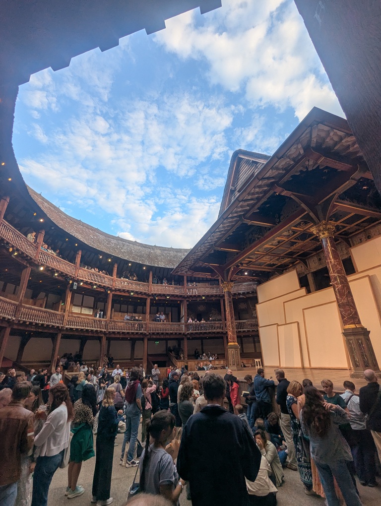 The interior of the Globe Theatre. People are standing in the ground area and some spectators have taken their seats. it is still light and the blue sky can be seen through the open roof, punctuated by white fluffy clouds