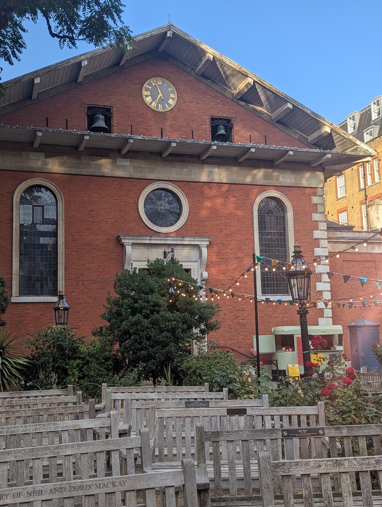 St Paul's Covent Garden. A red brick church with a gable end roof. Park benches are set out for the audience of a theatrical performance.